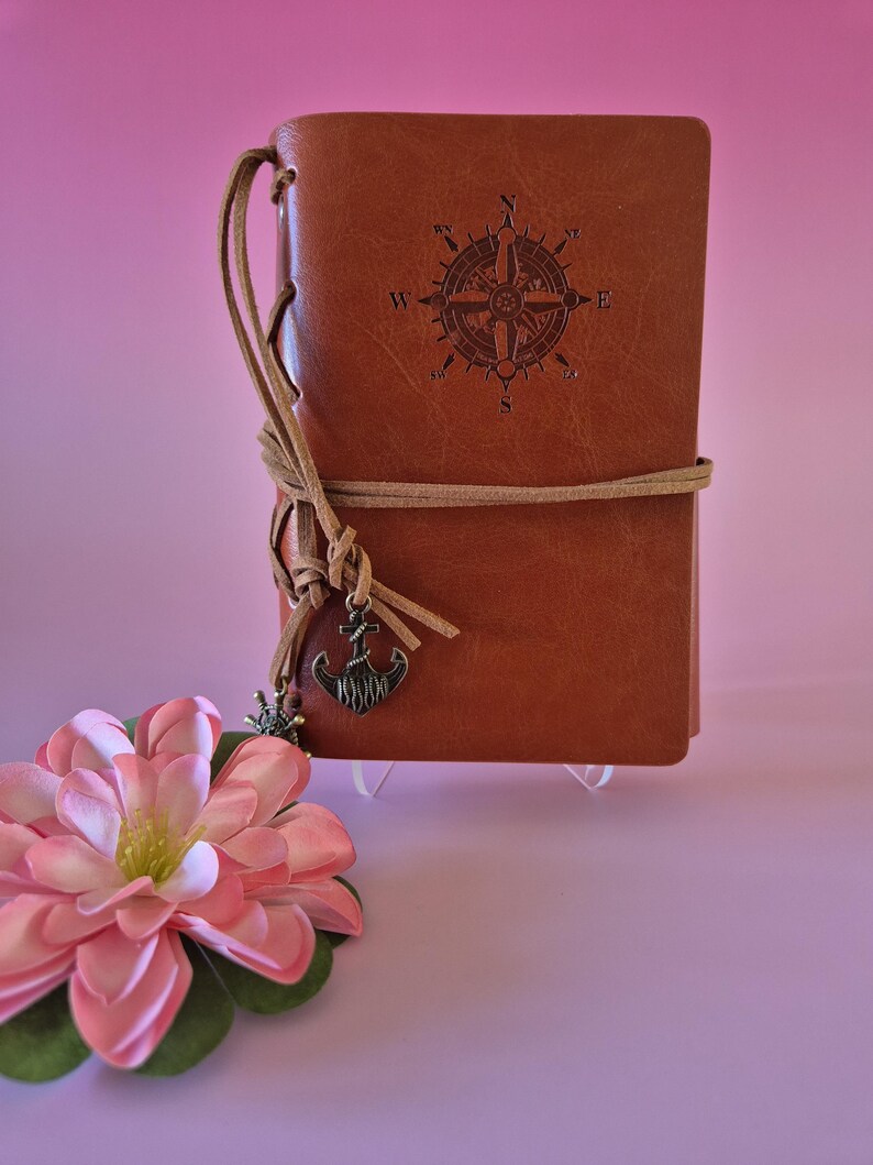 May include: A brown leather journal with a compass rose design embossed on the cover. The journal is tied with a tan leather cord and features a decorative anchor charm. A pink flower with green leaves is in the foreground.