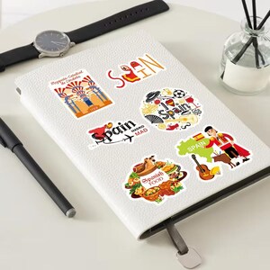 Pu&ograve; includere: Una collezione di adesivi a tema Spagna su un taccuino bianco. Gli adesivi presentano immagini di cibo spagnolo, un torero, una chitarra e la parola "Spagna" in vari disegni. Sono visibili anche una penna e un orologio neri.