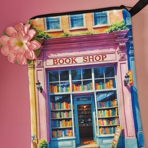 May include: A rectangular, colorful book shop design on a canvas bag. The bag features a pink and blue facade with the words "BOOK SHOP" above the entrance. The shop is filled with bookshelves and books. A pink flower is attached to the bag.