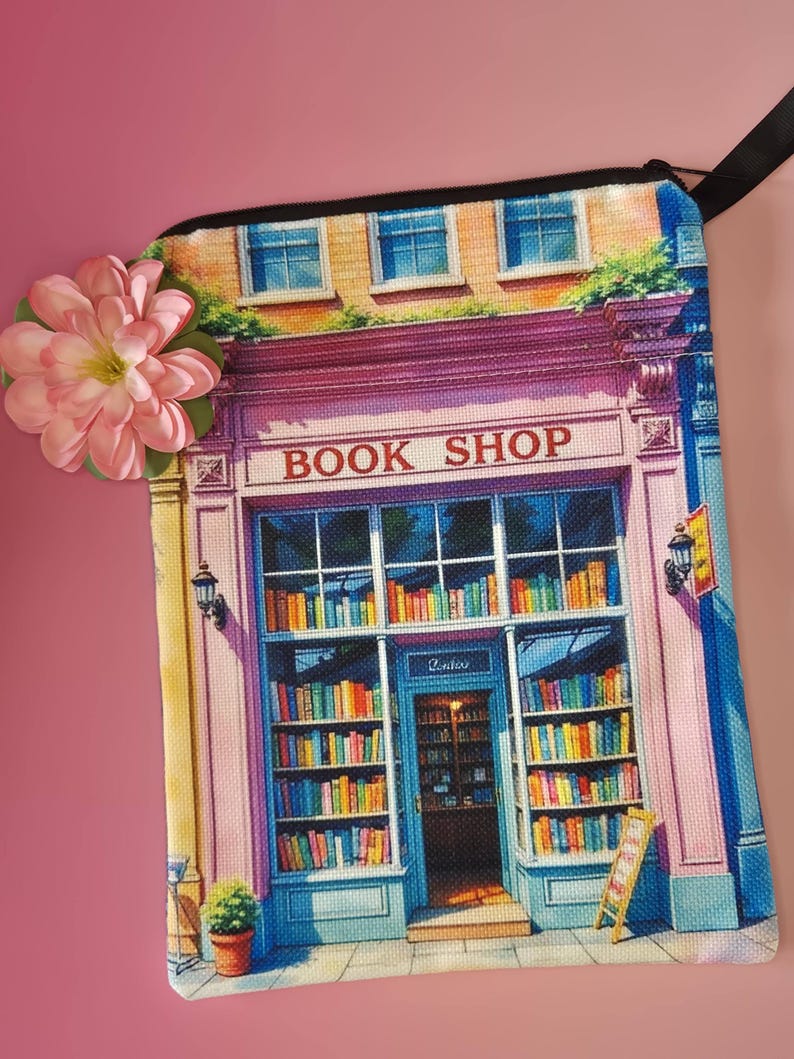 May include: A rectangular, colorful book shop design on a canvas bag. The bag features a pink and blue facade with the words "BOOK SHOP" above the entrance. The shop is filled with bookshelves and books. A pink flower is attached to the bag.