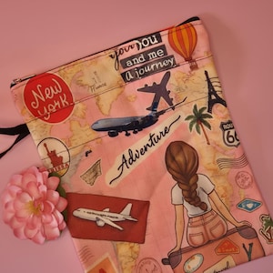 May include: Pink travel-themed pouch with a black zipper. The pouch features illustrations of landmarks, airplanes, and the text "New York", "Adventure", and "you and me a journey". A drawing of a person sitting on a suitcase is also visible.
