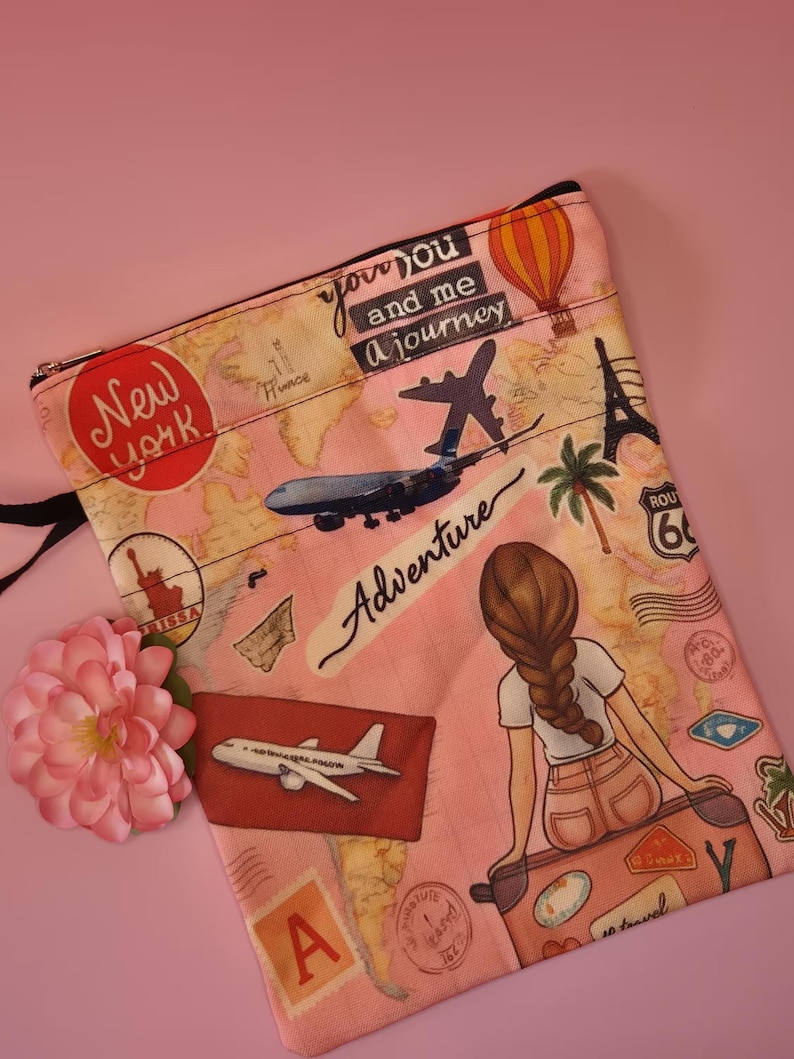 May include: Pink travel-themed pouch with a black zipper. The pouch features illustrations of landmarks, airplanes, and the text "New York", "Adventure", and "you and me a journey". A drawing of a person sitting on a suitcase is also visible.