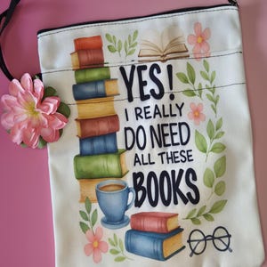 May include: A white canvas zippered pouch with a colorful book-themed design. The design includes stacks of books, a coffee cup, glasses, and the text "YES! I REALLY DO NEED ALL THESE BOOKS". A pink flower and black cord are attached.