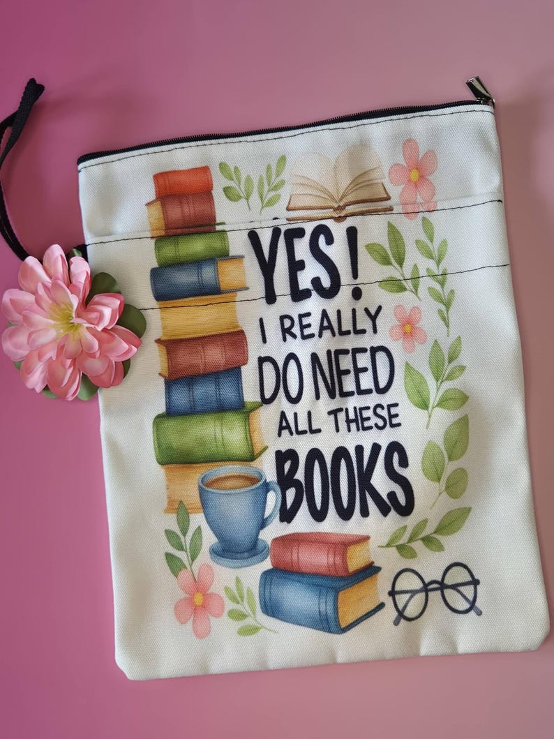 May include: A white canvas zippered pouch with a colorful book-themed design. The design includes stacks of books, a coffee cup, glasses, and the text "YES! I REALLY DO NEED ALL THESE BOOKS". A pink flower and black cord are attached.