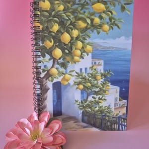 May include: A spiral-bound notebook featuring a vibrant illustration of a lemon tree laden with yellow fruit, overlooking a coastal scene with white buildings and blue water. A pink flower rests in the foreground.