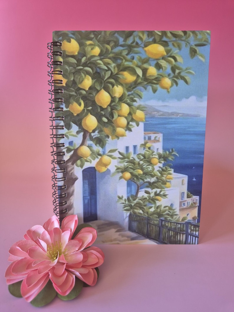 May include: A spiral-bound notebook featuring a vibrant illustration of a lemon tree laden with yellow fruit, overlooking a coastal scene with white buildings and blue water. A pink flower rests in the foreground.