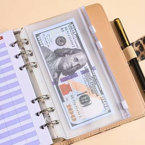 Op de afbeelding: Een beige ringbandplanner met een doorzichtige plastic zak met ritssluiting met een biljet van 100 dollar. Een zwarte pen met gouden accenten en een pennenhouder met luipaardprint zijn zichtbaar. De planner heeft gelinieerde pagina's.