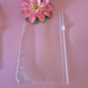 May include: A clear plastic binder pocket with a zip closure and three-hole punch. A pink and green artificial flower is placed on the top left corner. The background is a solid pink colour.