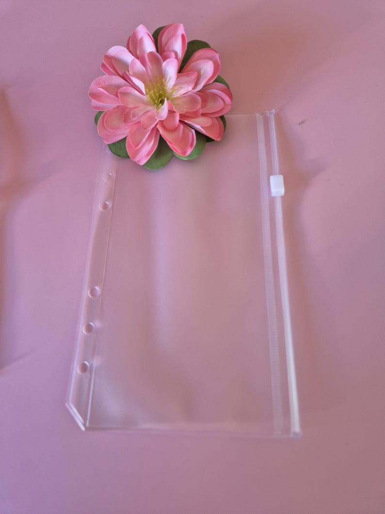 May include: A clear plastic binder pocket with a zip closure and three-hole punch. A pink and green artificial flower is placed on the top left corner. The background is a solid pink colour.