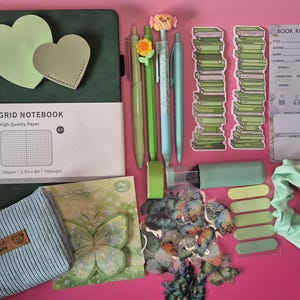 Pode incluir: Um conjunto de papelaria com um caderno de grelha A5 verde-escuro, quatro canetas com topos de flores e doces, dois marcadores de livros com pilhas de livros verdes, um cart&atilde;o de revis&atilde;o de livros, uma bolsa azul canelada, autocolantes de borboletas, fita adesiva verde e um el&aacute;stico de cabelo verde-claro.