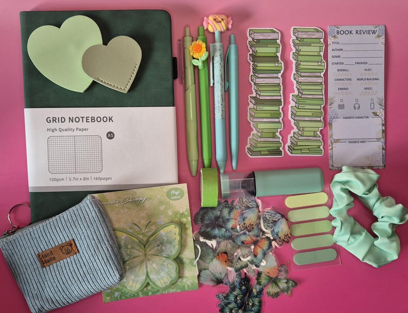 Pode incluir: Um conjunto de papelaria com um caderno de grelha A5 verde-escuro, quatro canetas com topos de flores e doces, dois marcadores de livros com pilhas de livros verdes, um cart&atilde;o de revis&atilde;o de livros, uma bolsa azul canelada, autocolantes de borboletas, fita adesiva verde e um el&aacute;stico de cabelo verde-claro.