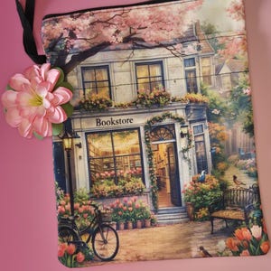 May include: A rectangular, zippered pouch featuring a detailed illustration of a bookstore with the word "Bookstore" above the entrance. The scene includes a bicycle, flowers, and a bench, with a pink flower attached to the black strap.