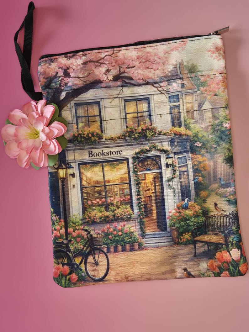 May include: A rectangular, zippered pouch featuring a detailed illustration of a bookstore with the word "Bookstore" above the entrance. The scene includes a bicycle, flowers, and a bench, with a pink flower attached to the black strap.