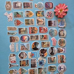May include: A collection of travel-themed stickers featuring landmarks, compasses, and the word "travel." The stickers showcase various destinations like London, Paris, and Italy, with a pink flower in the upper right corner. The stickers are on a blue background.