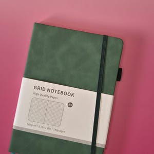 May include: A dark green A5 grid notebook with a black elastic closure band and a white paper belly band. The belly band features the text GRID NOTEBOOK, High Quality Paper, A5, 100gsm, 14.5cm x 20.3cm, and 160pages, along with a grid pattern graphic.