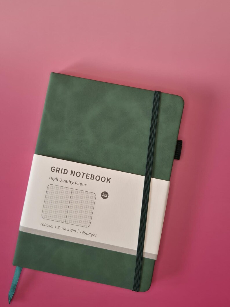 May include: A dark green A5 grid notebook with a black elastic closure band and a white paper belly band. The belly band features the text GRID NOTEBOOK, High Quality Paper, A5, 100gsm, 14.5cm x 20.3cm, and 160pages, along with a grid pattern graphic.