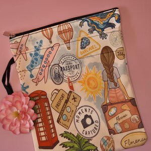 May include: A travel-themed zippered pouch with a white background, featuring illustrations of airplanes, hot air balloons, passport stamps, a red telephone booth, and a girl sitting on a suitcase. The pouch has a black zipper and a black loop.