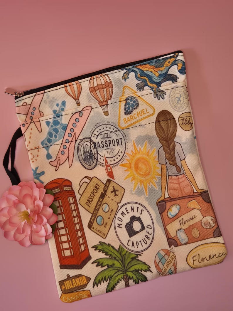 May include: A travel-themed zippered pouch with a white background, featuring illustrations of airplanes, hot air balloons, passport stamps, a red telephone booth, and a girl sitting on a suitcase. The pouch has a black zipper and a black loop.