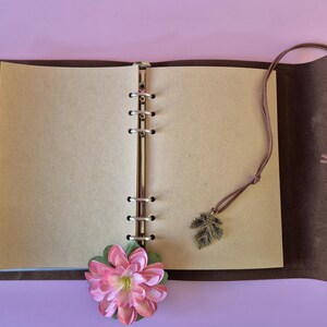 May include: Open brown leather journal with blank pages. A pink and green flower and a leaf charm dangle from the journal. The journal is open on a light purple surface.