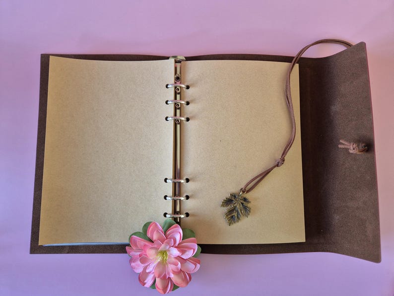 May include: Open brown leather journal with blank pages. A pink and green flower and a leaf charm dangle from the journal. The journal is open on a light purple surface.