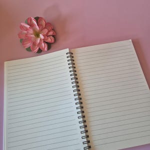May include: An open, lined notebook with a black spiral binding rests on a pink surface. A pink and green artificial flower is positioned near the notebook. The notebook's pages are blank and ready for writing.