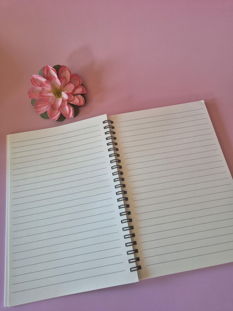 May include: An open, lined notebook with a black spiral binding rests on a pink surface. A pink and green artificial flower is positioned near the notebook. The notebook's pages are blank and ready for writing.