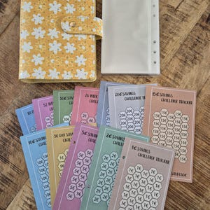 K&ouml;nnte beinhalten: Ein gelber Planer mit Blumenmuster und Druckknopfverschluss, ein transparenter Ordner-Einsatz und mehrere farbenfrohe Spar-Challenge-Tracker. Die Tracker zeigen Betr&auml;ge von 1&euro; bis 100&euro;, ideal f&uuml;r die Budgetplanung.
