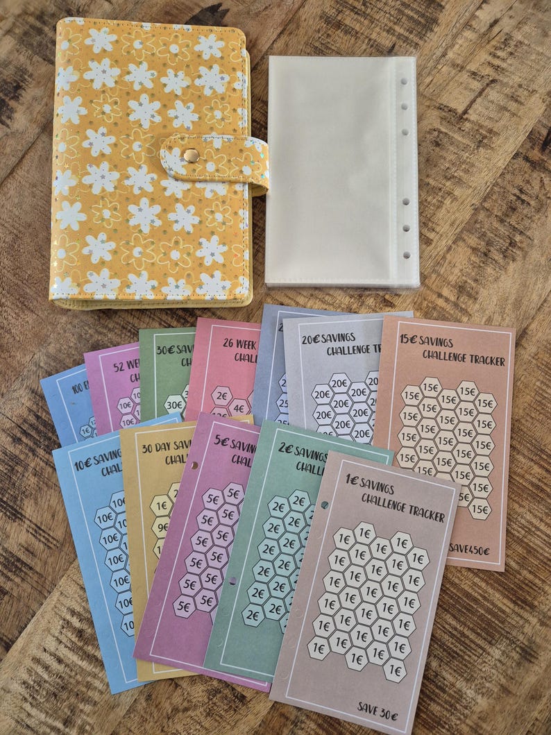 K&ouml;nnte beinhalten: Ein gelber Planer mit Blumenmuster und Druckknopfverschluss, ein transparenter Ordner-Einsatz und mehrere farbenfrohe Spar-Challenge-Tracker. Die Tracker zeigen Betr&auml;ge von 1&euro; bis 100&euro;, ideal f&uuml;r die Budgetplanung.