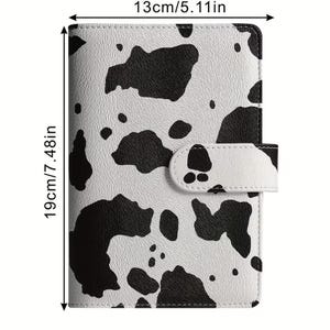 Op de afbeelding: Een planner met een koeienprint, 13 cm breed en 19 cm hoog. De omslag heeft een patroon van zwarte vlekken op een witte achtergrond. Het heeft een magnetische sluiting en een gestructureerd oppervlak.