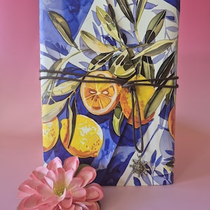 Op de afbeelding: Een notitieboek met een aquarelontwerp van citroenen en olijftakken, gebonden met een bruin koord. De omslag heeft een blauw-wit patroon. Een kleine hanger hangt aan het koord. Een roze bloem staat op de voorgrond.