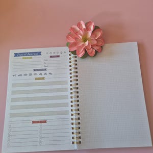 Op de afbeelding: Open reisdagboek met een roze bloem op een roze achtergrond. Het dagboek heeft een spiraalbinding en een checklistgedeelte. De omslag luidt "Travel Journal". De pagina's zijn wit met gestippelde rasterlijnen.