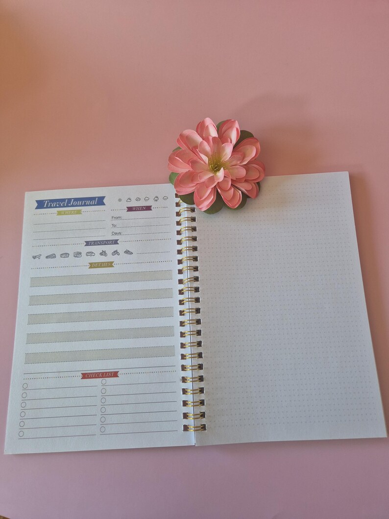Op de afbeelding: Open reisdagboek met een roze bloem op een roze achtergrond. Het dagboek heeft een spiraalbinding en een checklistgedeelte. De omslag luidt "Travel Journal". De pagina's zijn wit met gestippelde rasterlijnen.
