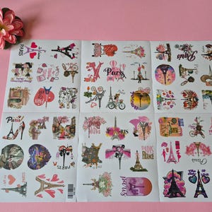 May include: Sheets of stickers with a Paris theme, featuring the Eiffel Tower, bicycles, and floral designs. The stickers are in various shapes and sizes, with pink, red, and black accents. A pink flower is in the upper left corner.