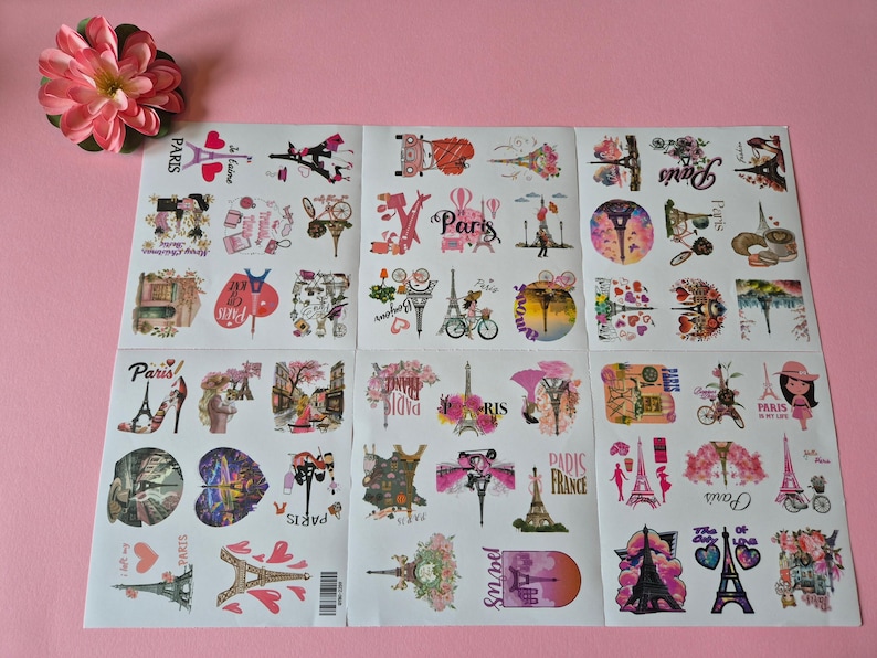 May include: Sheets of stickers with a Paris theme, featuring the Eiffel Tower, bicycles, and floral designs. The stickers are in various shapes and sizes, with pink, red, and black accents. A pink flower is in the upper left corner.