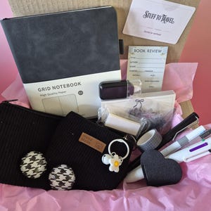 Puede incluir: Una caja de regalo de papeler&iacute;a que contiene un cuaderno de cuadr&iacute;cula A5 negro, dos estuches acanalados negros con botones de pata de gallo, una tarjeta Stuff to Travel, una ficha de lectura, un recipiente negro, un clip de coraz&oacute;n, bol&iacute;grafos y cinta washi.