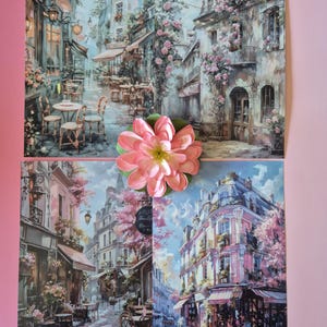以下が含まれることがあります： カフェや花のアクセントがあるヨーロッパの街並みを描いた4つのアートプリント。大きなピンクの花がプリントの上に置かれています。プリントには、日よけ、テーブル、椅子のある建物が描かれており、ロマンチックな雰囲気を作り出しています。