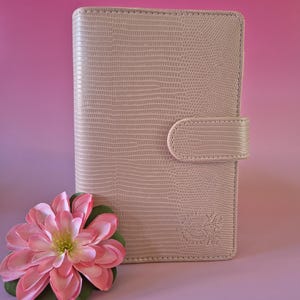 Pu&ograve; includere: Un diario beige chiaro, strutturato, con chiusura a scatto. Un fiore rosa e verde &egrave; in primo piano. Il diario ha un logo in rilievo nell'angolo in basso a destra. Lo sfondo &egrave; una sfumatura di rosa e viola.