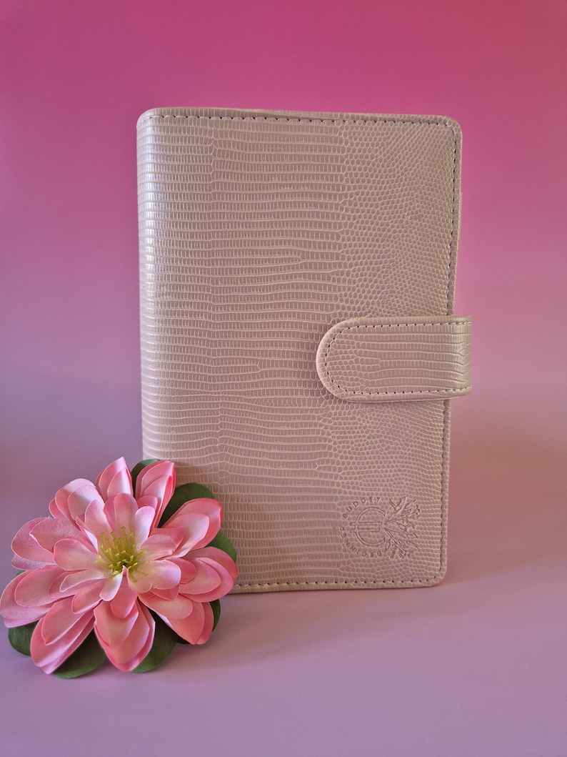 Pu&ograve; includere: Un diario beige chiaro, strutturato, con chiusura a scatto. Un fiore rosa e verde &egrave; in primo piano. Il diario ha un logo in rilievo nell'angolo in basso a destra. Lo sfondo &egrave; una sfumatura di rosa e viola.