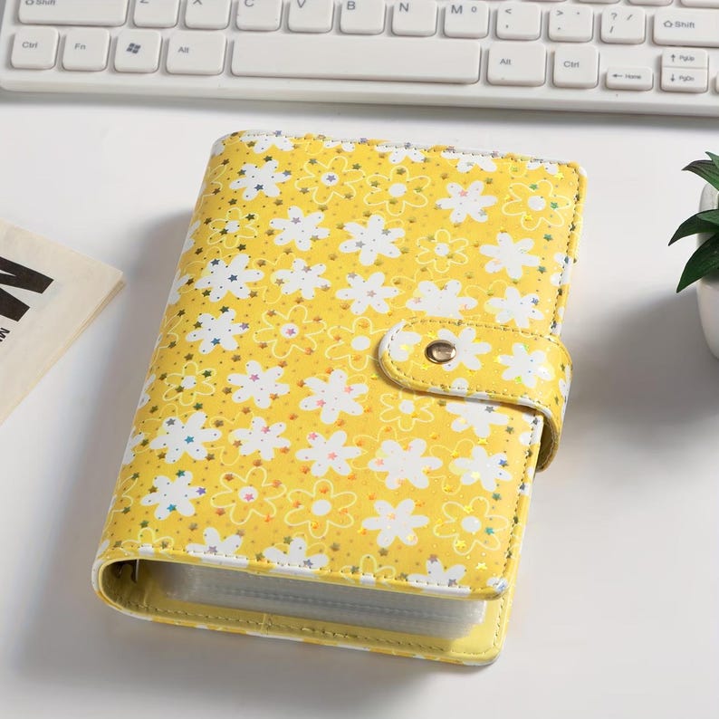 K&ouml;nnte beinhalten: Ein gelber Ordner mit floralem Muster und Druckknopfverschluss. Das Design zeigt wei&szlig;e Blumen mit gelben Zentren. Der Ordner ist geschlossen und scheint aus einem lederartigen Material zu bestehen.