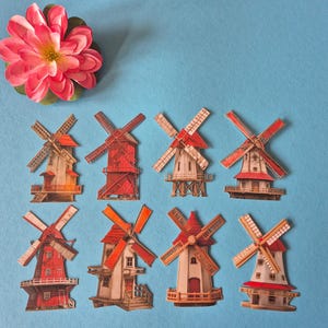 May include: A collection of miniature windmill cutouts with red and white accents, arranged on a blue background. A pink and yellow flower is in the upper left corner. The windmills vary in design and color.