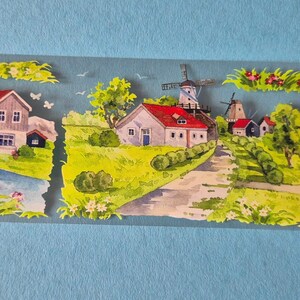 Op de afbeelding: Decoratieve washi tape met een schilderachtig landschap met windmolens, huizen en een rivier. De tape heeft een lichtblauwe achtergrond en kleurrijke illustraties van landelijke taferelen met roodbedekte gebouwen en groene velden.