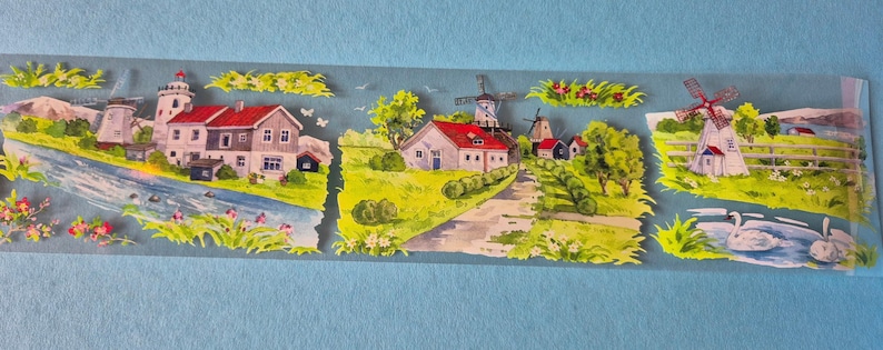 Op de afbeelding: Decoratieve washi tape met een schilderachtig landschap met windmolens, huizen en een rivier. De tape heeft een lichtblauwe achtergrond en kleurrijke illustraties van landelijke taferelen met roodbedekte gebouwen en groene velden.
