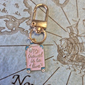 Puede incluir: Un cierre giratorio dorado con un dije de maleta rosa. La maleta tiene ribetes azules y las palabras "To travel is to live" en rosa. El dije tiene peque&ntilde;as ruedas blancas, sobre un fondo de mapa.