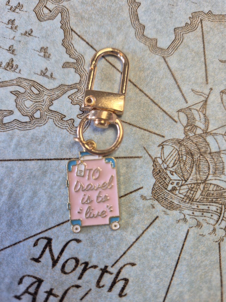 Puede incluir: Un cierre giratorio dorado con un dije de maleta rosa. La maleta tiene ribetes azules y las palabras "To travel is to live" en rosa. El dije tiene peque&ntilde;as ruedas blancas, sobre un fondo de mapa.