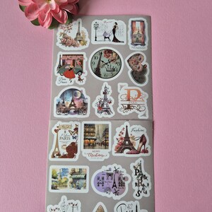 May include: A sheet of Paris-themed stickers featuring the Eiffel Tower, fashion illustrations, and vintage clock designs. The stickers include the text "Paris", "Fashion", and "Merry Christmas". A pink flower is in the upper left corner.