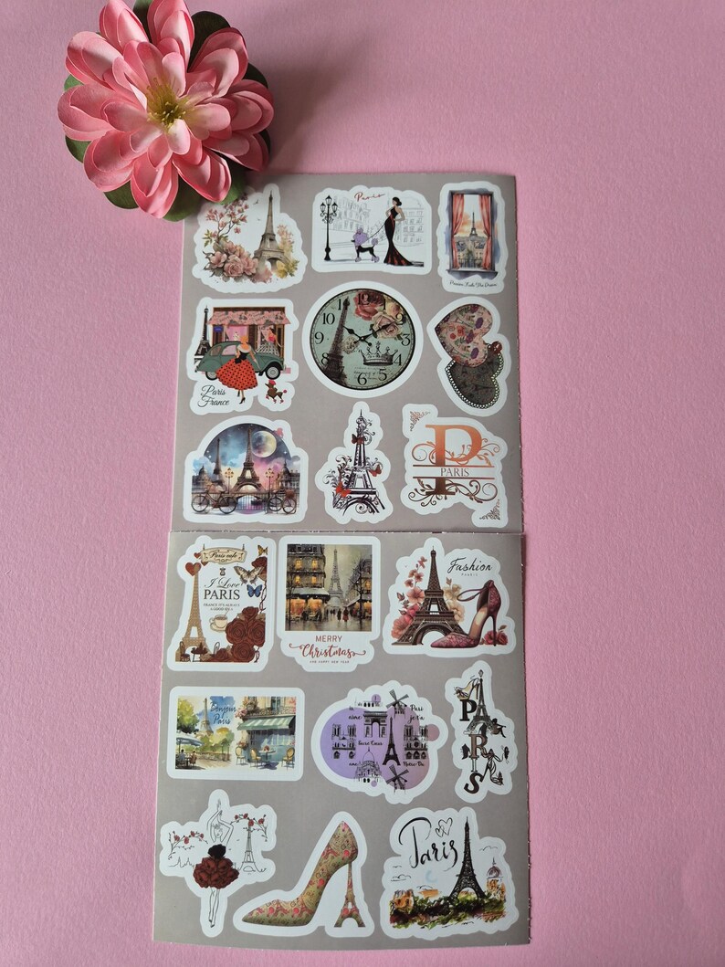 May include: A sheet of Paris-themed stickers featuring the Eiffel Tower, fashion illustrations, and vintage clock designs. The stickers include the text "Paris", "Fashion", and "Merry Christmas". A pink flower is in the upper left corner.