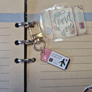 Puede incluir: Un llavero dorado con un dije en forma de boleto rosa y blanco que dice "Adventure Awaits" y presenta un peque&ntilde;o gr&aacute;fico de avi&oacute;n. El llavero est&aacute; unido a un cuaderno con una pegatina con tem&aacute;tica de viajes que dice "Travel the World".