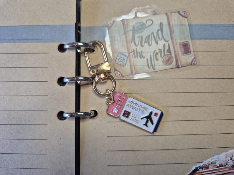 Puede incluir: Un llavero dorado con un dije en forma de boleto rosa y blanco que dice "Adventure Awaits" y presenta un peque&ntilde;o gr&aacute;fico de avi&oacute;n. El llavero est&aacute; unido a un cuaderno con una pegatina con tem&aacute;tica de viajes que dice "Travel the World".