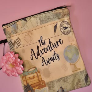 May include: A beige travel-themed pouch with a black zipper, featuring a vintage map design and the words "The Adventure Awaits." The pouch includes illustrations of luggage, a globe, and a camera. A pink flower is placed on the side.