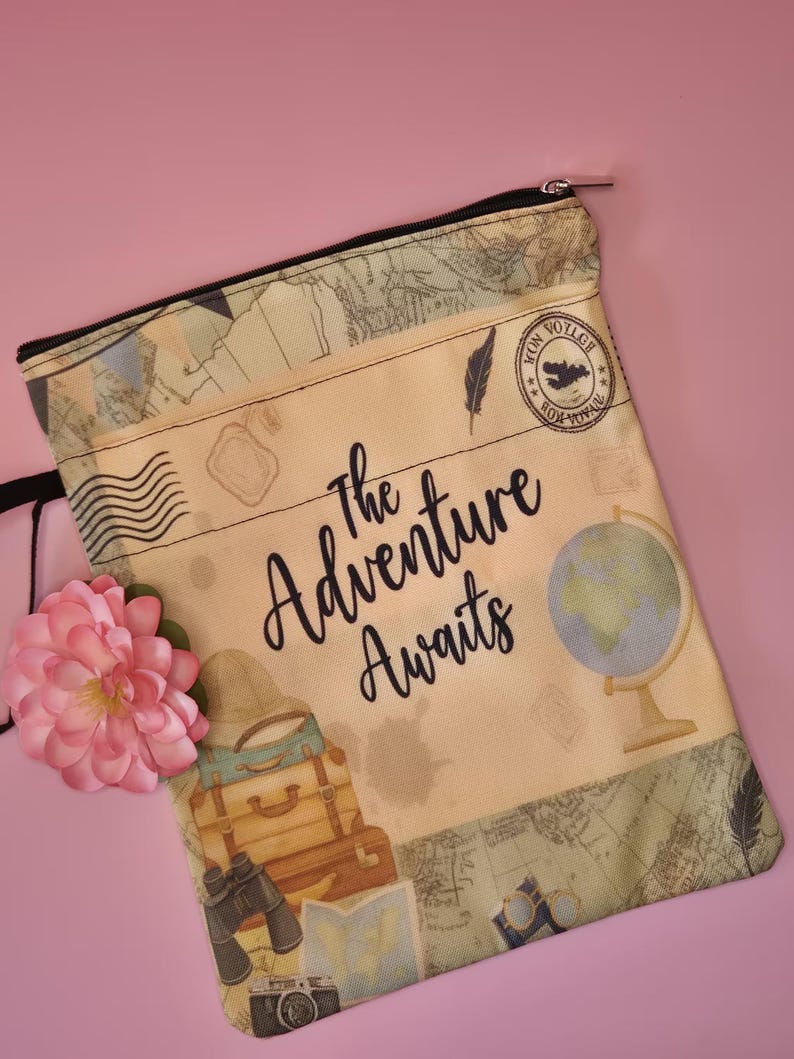 May include: A beige travel-themed pouch with a black zipper, featuring a vintage map design and the words "The Adventure Awaits." The pouch includes illustrations of luggage, a globe, and a camera. A pink flower is placed on the side.